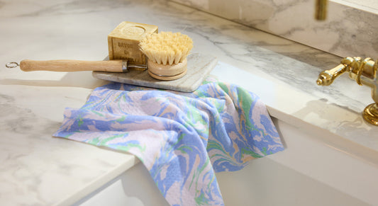 A wooden dish brush, a bar of soap, and a patterned blue and purple cloth rest on a marble countertop above a white sink with a brass faucet.