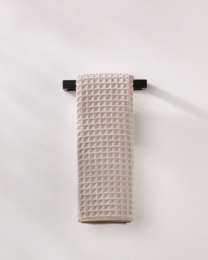 A GEOMETRY Taupe waffle hand towel made from recycled materials hangs neatly on a sleek black towel bar against a plain white wall.