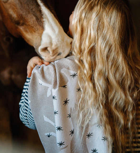 A woman with long, wavy blonde hair gently nuzzles a brown horse with a white stripe on its face. They share an affectionate moment indoors.